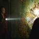 Tess and Joel look at a mushroom man stuck to the wall.